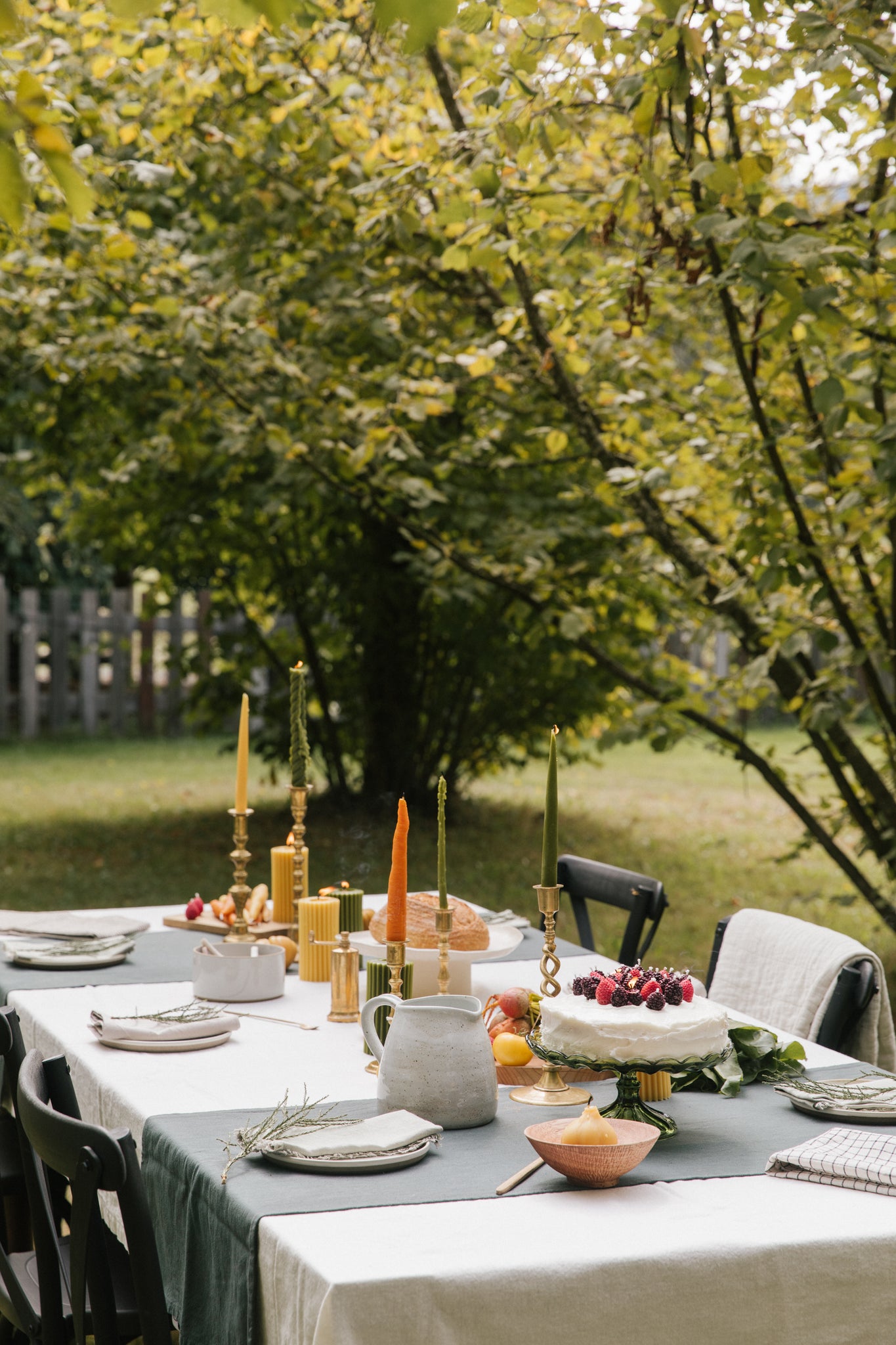 An outdoor dining table set in a garden with white tablecloths, dark green runners, brass candlesticks with yellow and green taper candles, plates, a small cake, and bowls of fruit, surrounded by trees.