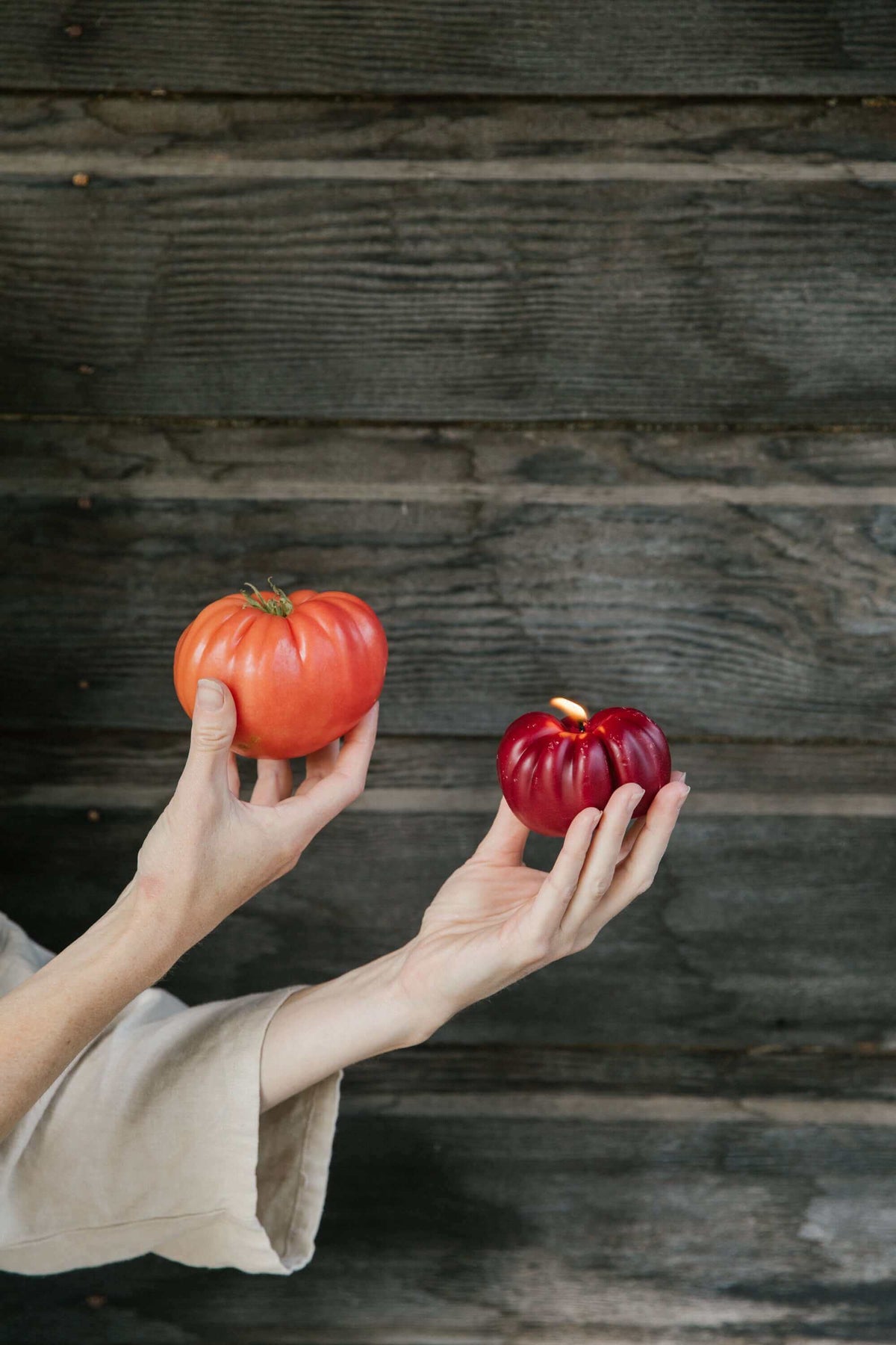 Person holding heirloom tomato candle in sun kissed orange and heirloom tomatoes against rustic wooden background.