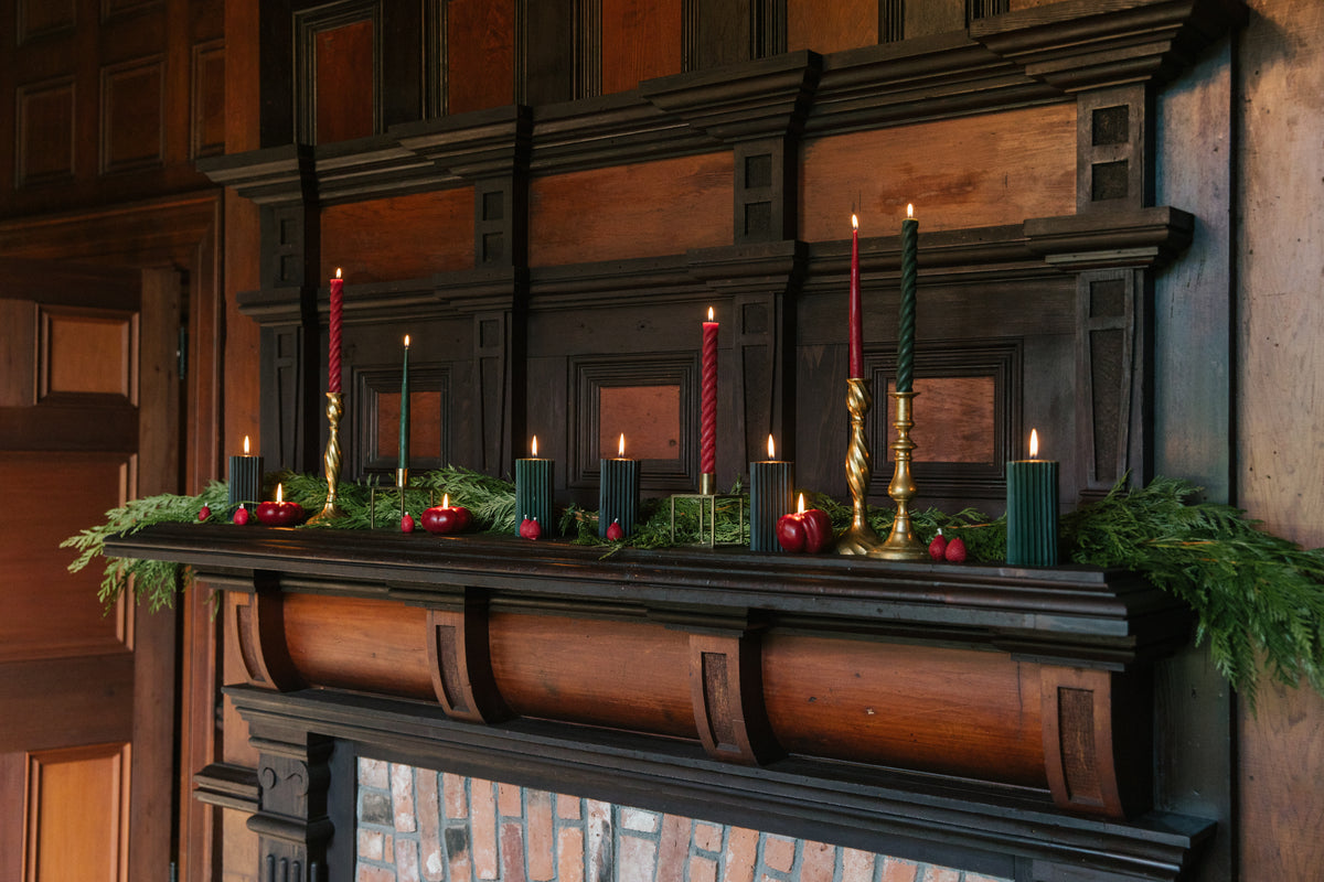 Red and green beeswax candles lit among evergreen branches on a dark wood fireplace mantel.