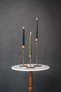 Green beeswax taper candles in raw silver candlesticks on a small marble table against a black background