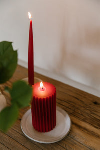 A red beeswax pillar candle sitting on a ceramic dish next to a red beeswax taper candle both on a rustic wooden table with greenery peeking into the frame