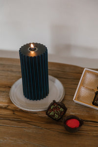 Dark blue beeswax pillar candle burning on a ceramic plate with festive holiday chocolate next to it