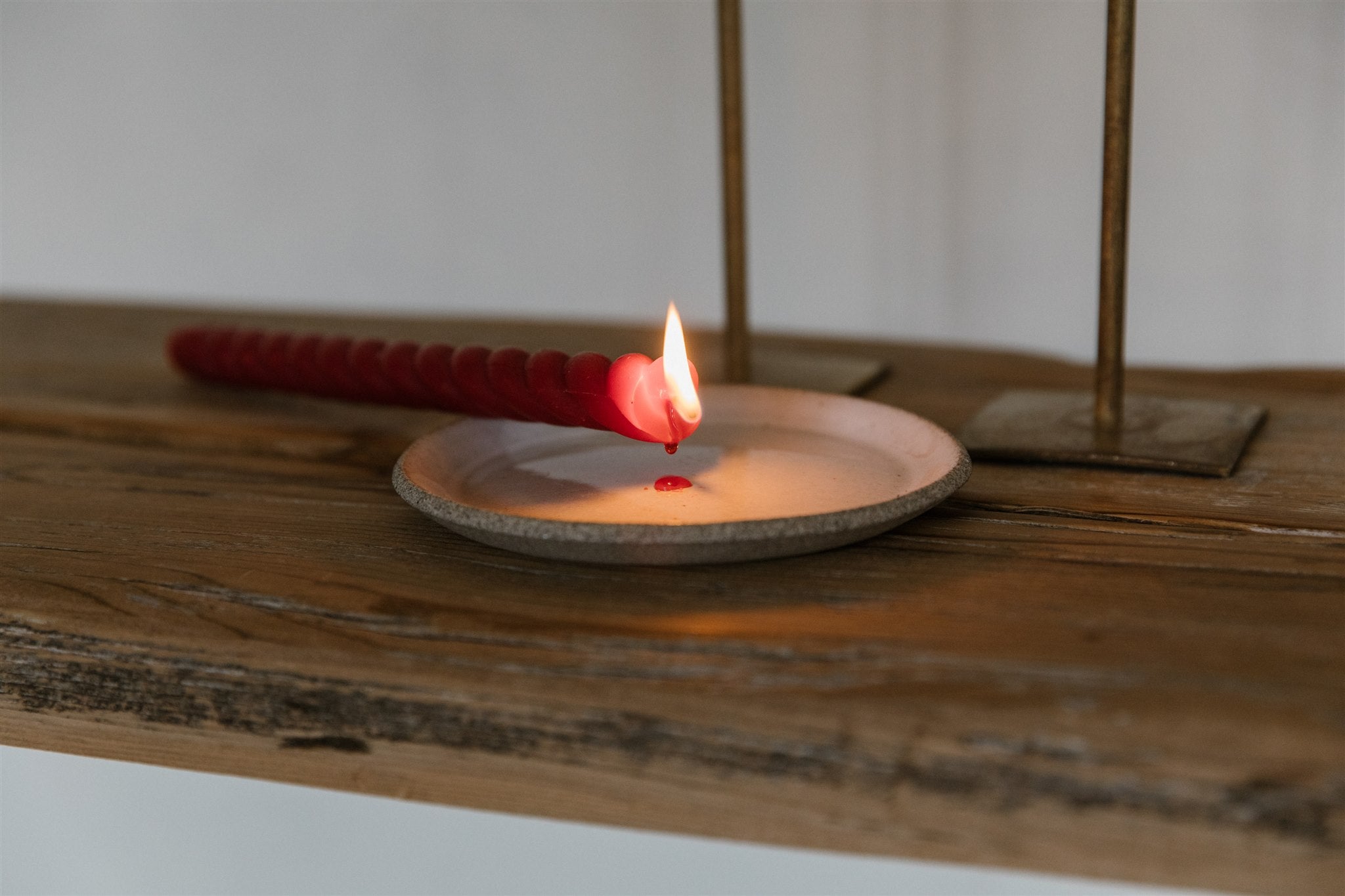 Close up of red twisted beeswax taper candle on its side dripping onto a ceramic plate sitting on a rustic wooden table