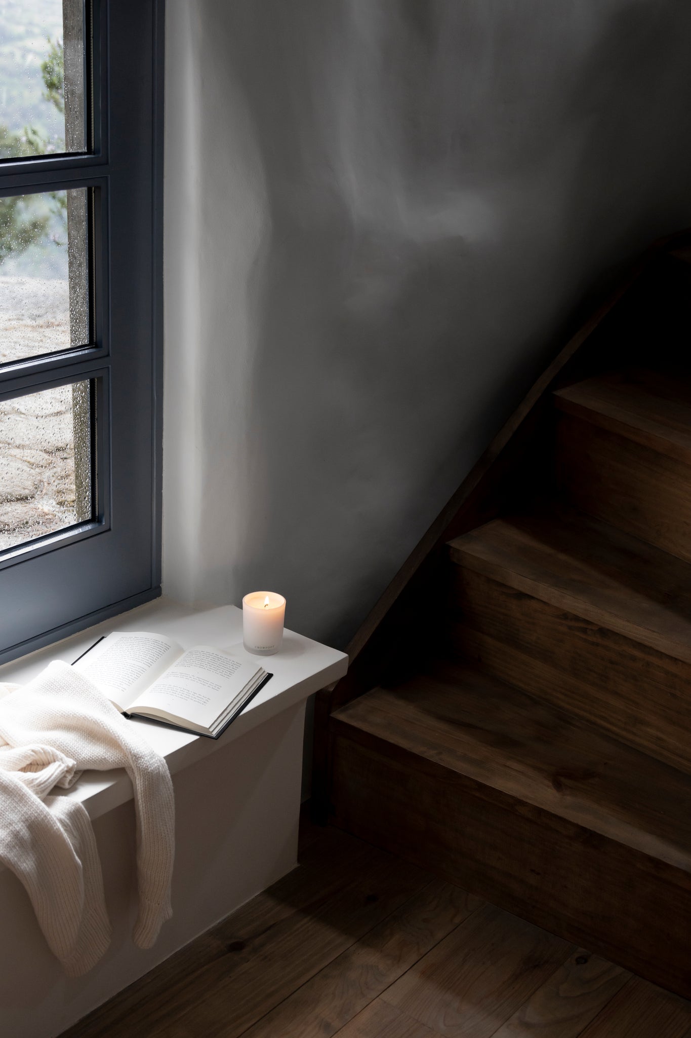Lit Crowfoot Collective candle beside an open book and blanket by a sunlit staircase.