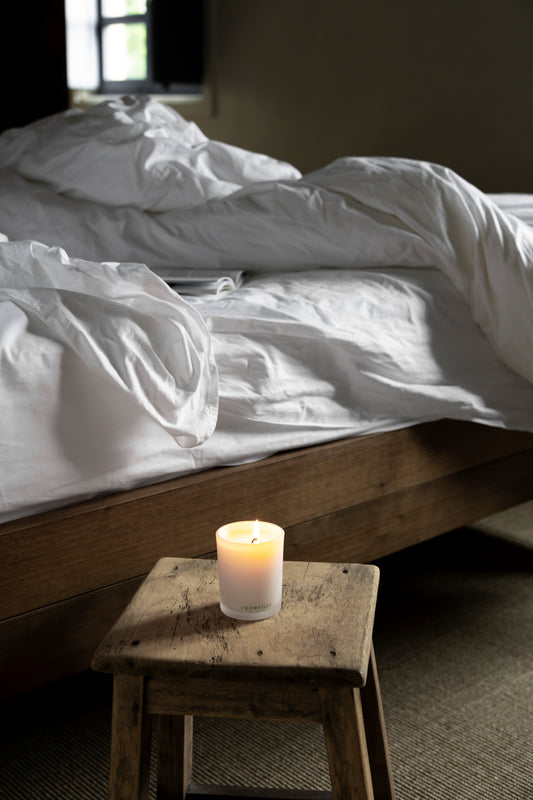 Lit candle on a wooden stool beside a linen bed